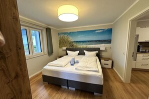 Un dormitorio con cama, una mesita de noche, un cuadro de una playa y una ventana.