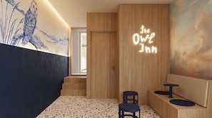 Un hall de hotel con un mostrador de recepción de madera, dos zonas de estar, un mural y un letrero de neón que dice 'The Owl Inn'.