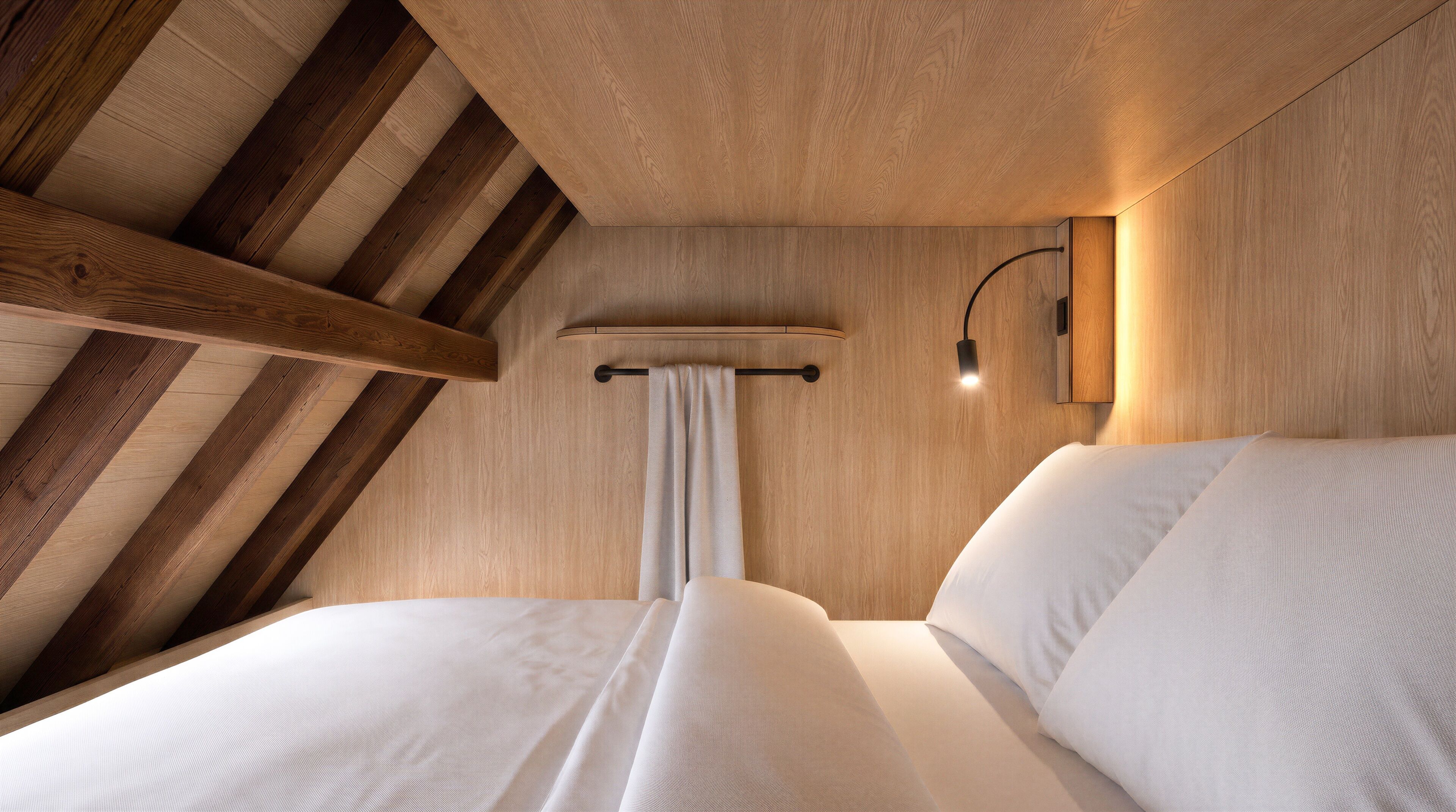 A room with a sloped ceiling, wooden beams, a bed with white bedding, a wall-mounted light, and a curtain.