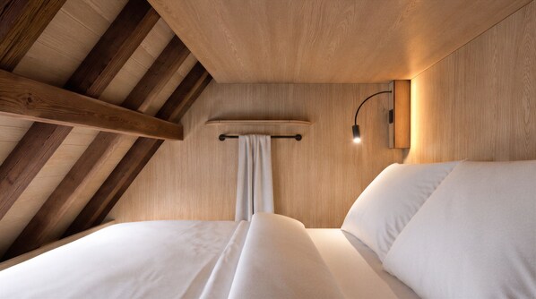 A room with a sloped ceiling, wooden beams, a bed with white bedding, a wall-mounted light, and a curtain.