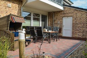 Een terras met een tafel en stoelen, een rieten stoel met een gestreept kussen, en een witte deur.