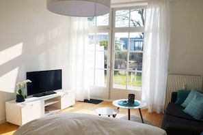 A modern hotel room with a bed, a TV on a white console, a small round table, and a view of the outdoors through large windows. - Apartment in Travemünde with swimming pool (Travemünde)