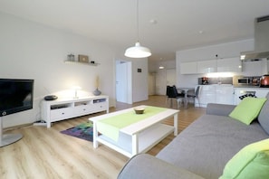 A modern living room with a flat-screen TV, white furniture, a dining area, and a kitchen. - Holiday apartment with 1 bedroom (Dierhagen)