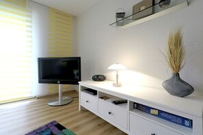 A modern living room with a flat-screen TV, a lamp, a vase with dried plants, and a shelf with decorative items. - Holiday apartment with 1 bedroom (Dierhagen)