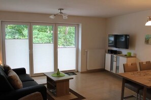 A living room with a sofa, a coffee table, a dining table with chairs, a television, and a sliding glass door leading to a balcony. - Holiday apartment with 2 bedrooms (Norddeich)