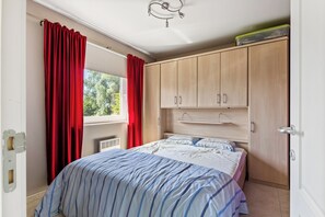 A bedroom with a bed, red curtains, wooden wardrobe, and a window with a view of trees. - Green Garden 9 / 0105 (De Haan)
