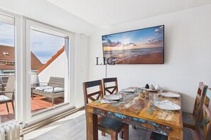A dining area with a wooden table, chairs, and a television mounted on the wall displaying a beach scene. - Holiday apartment with 2 bedrooms (Langeoog)