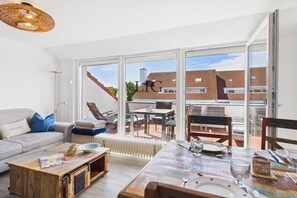 A modern living room with a sofa, coffee table, and dining table set for two. Large windows offer a view of the neighborhood. - Holiday apartment with 2 bedrooms (Langeoog)