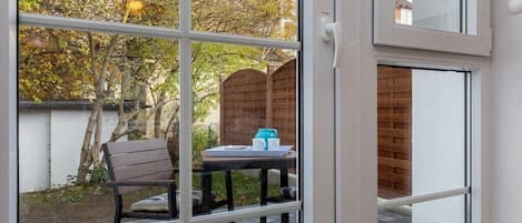 Vista a través de una ventana que muestra un patio con una valla de madera, una mesa con una silla y un muro de ladrillo.