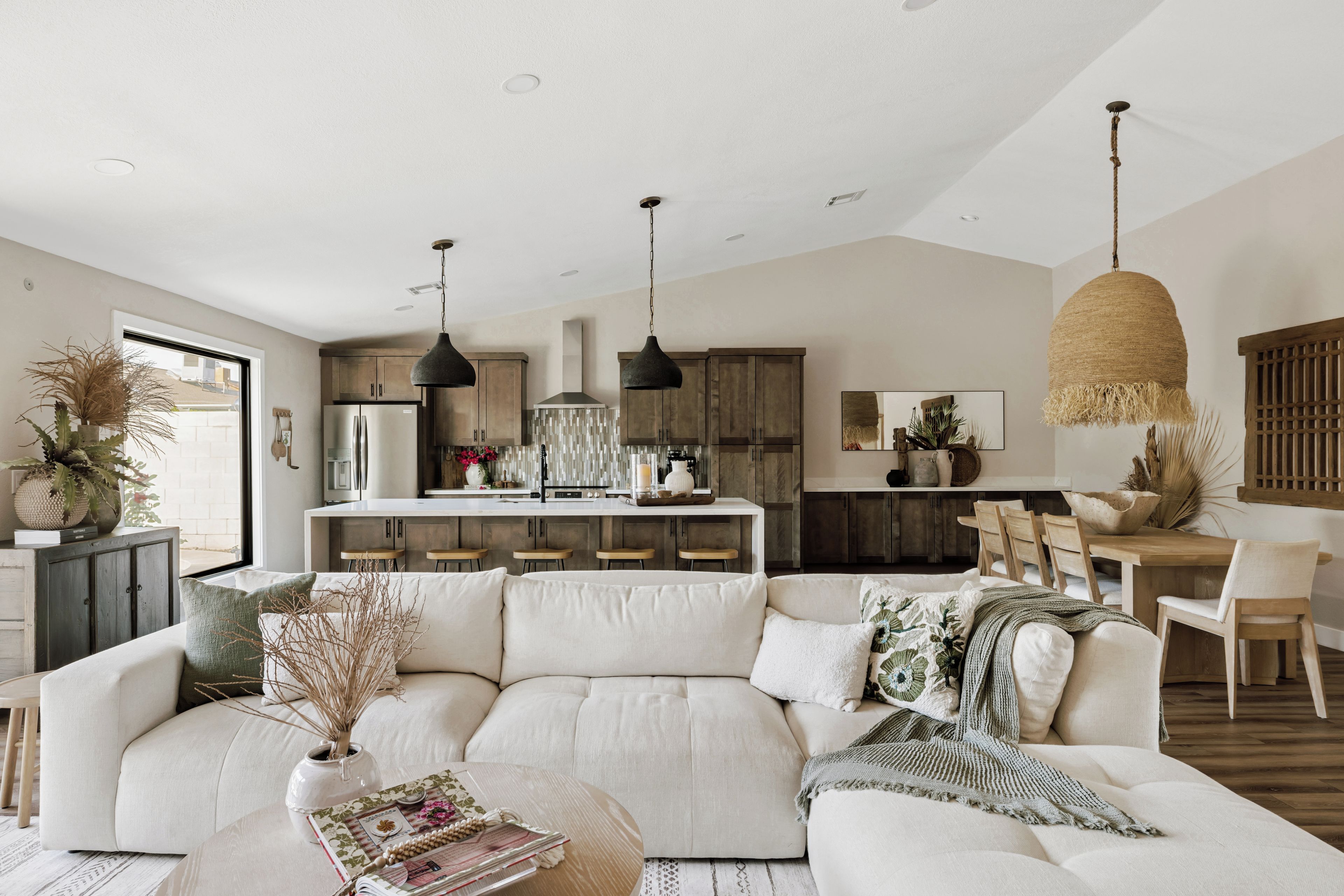 A modern living room with a sectional sofa, a dining area with wooden chairs, and a kitchen with pendant lighting.