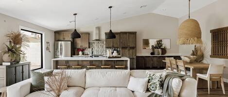 A modern living room with a sectional sofa, a dining area with wooden chairs, and a kitchen with pendant lighting.