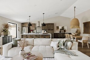 A modern living room with a sectional sofa, a dining area with wooden chairs, and a kitchen with pendant lighting.