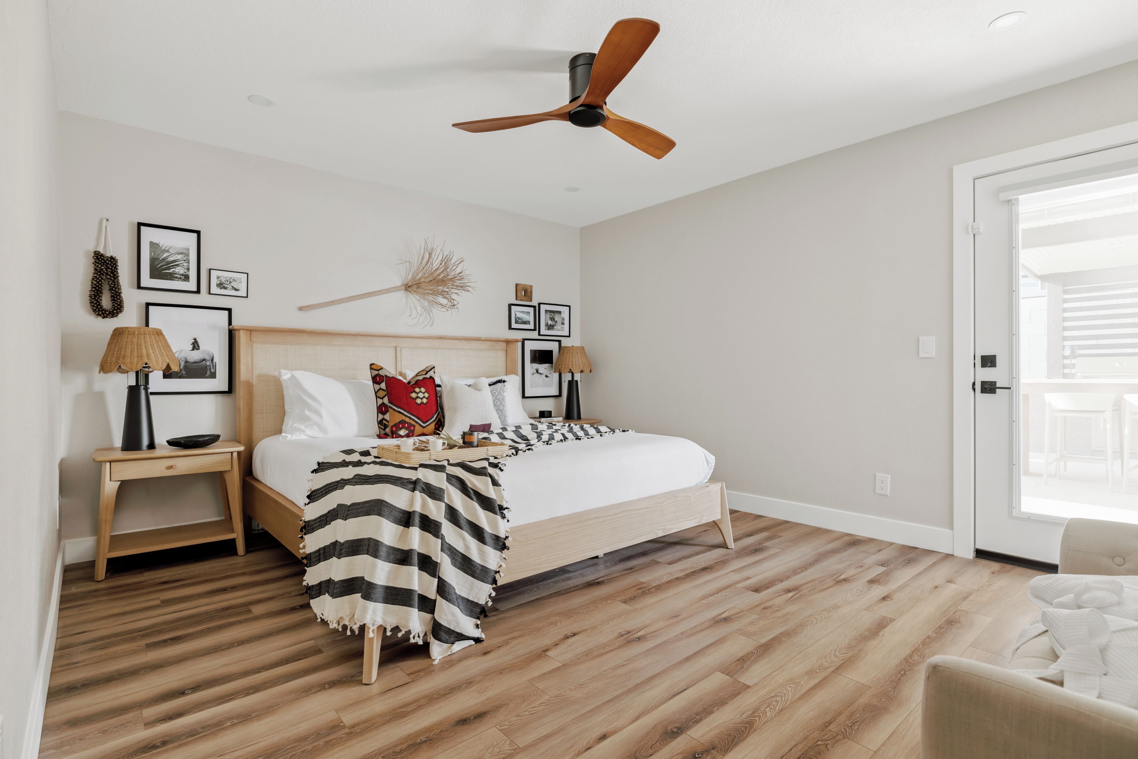 Une chambre à coucher avec un lit, des tables de chevet, un ventilateur de plafond et une vue sur l’extérieur, visible à travers une porte.