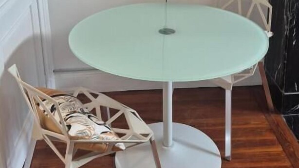 Une table ronde avec un pied blanc, une seule chaise dotée d’un siège en osier, et un luminaire fixé au mur.