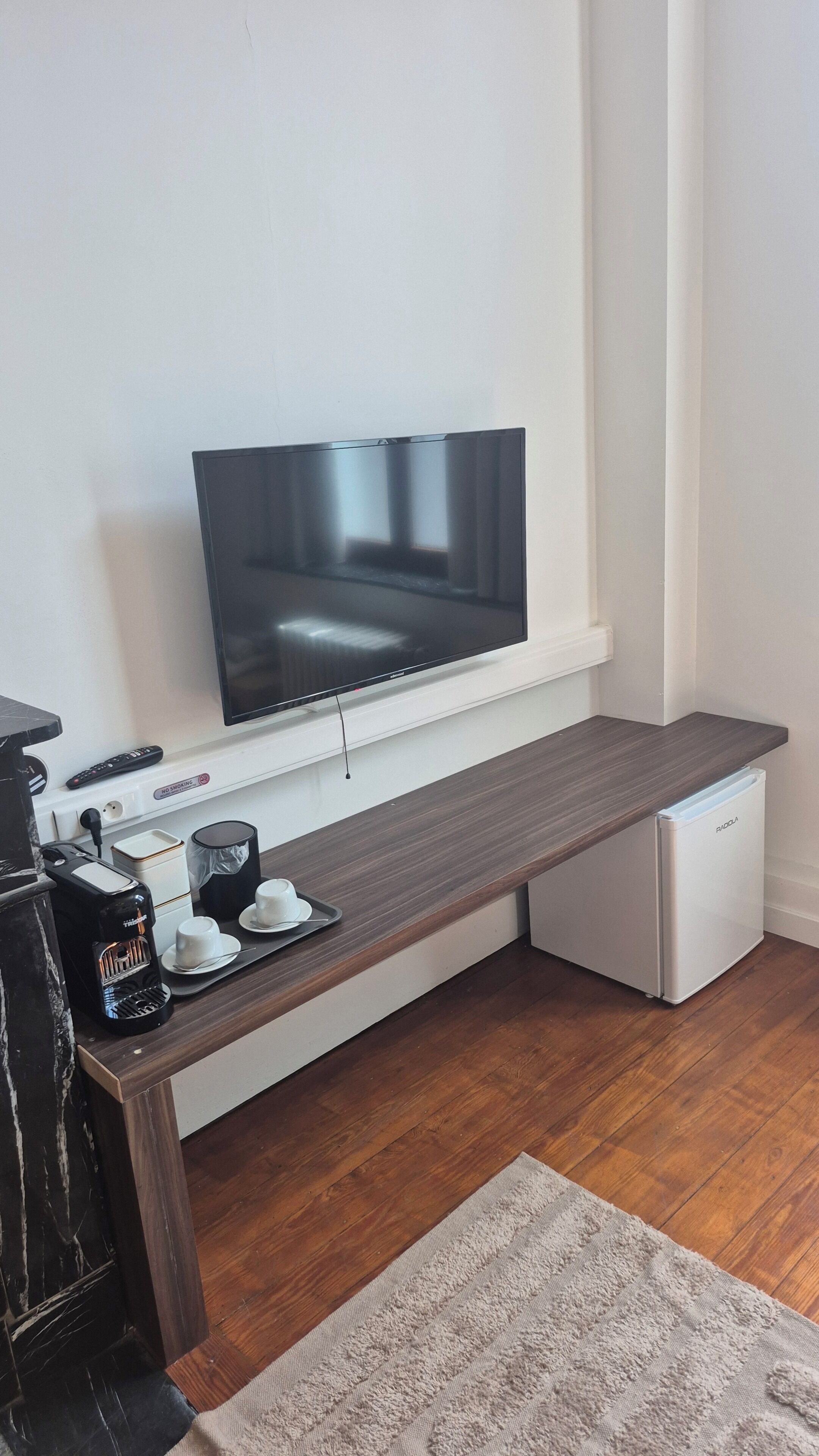 Een moderne woonruimte met een flatscreen televisie aan een witte muur, een houten bank met een koffiezetapparaat en kopjes, en een compacte koelkast.