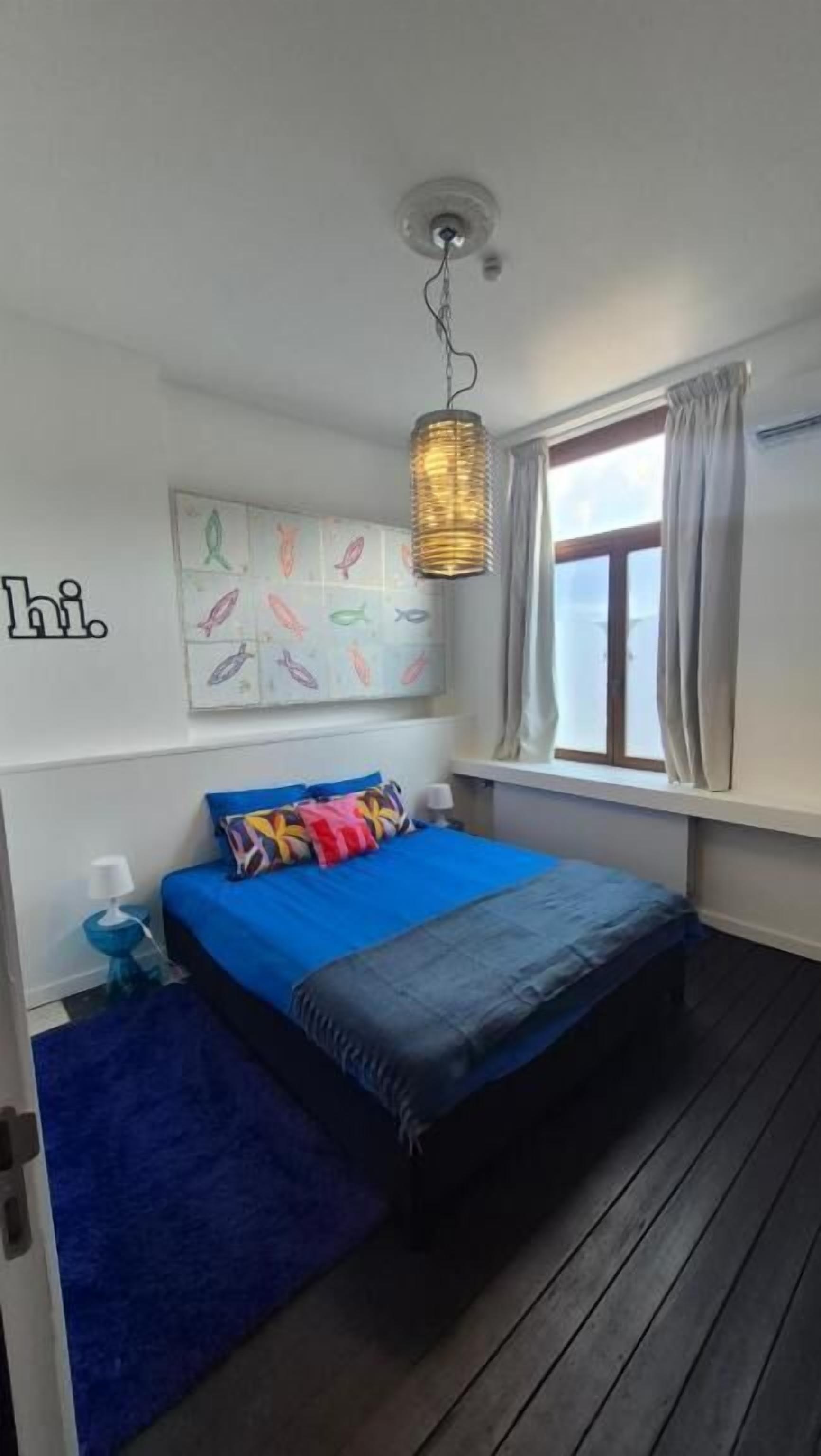 A bedroom with a bed, a blue rug, a wall with artwork, a hanging light fixture, and a window with curtains.
