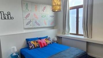 Un dormitorio con cama, un tapete azul, una pared con obras de arte, una lámpara colgante y una ventana con cortinas.