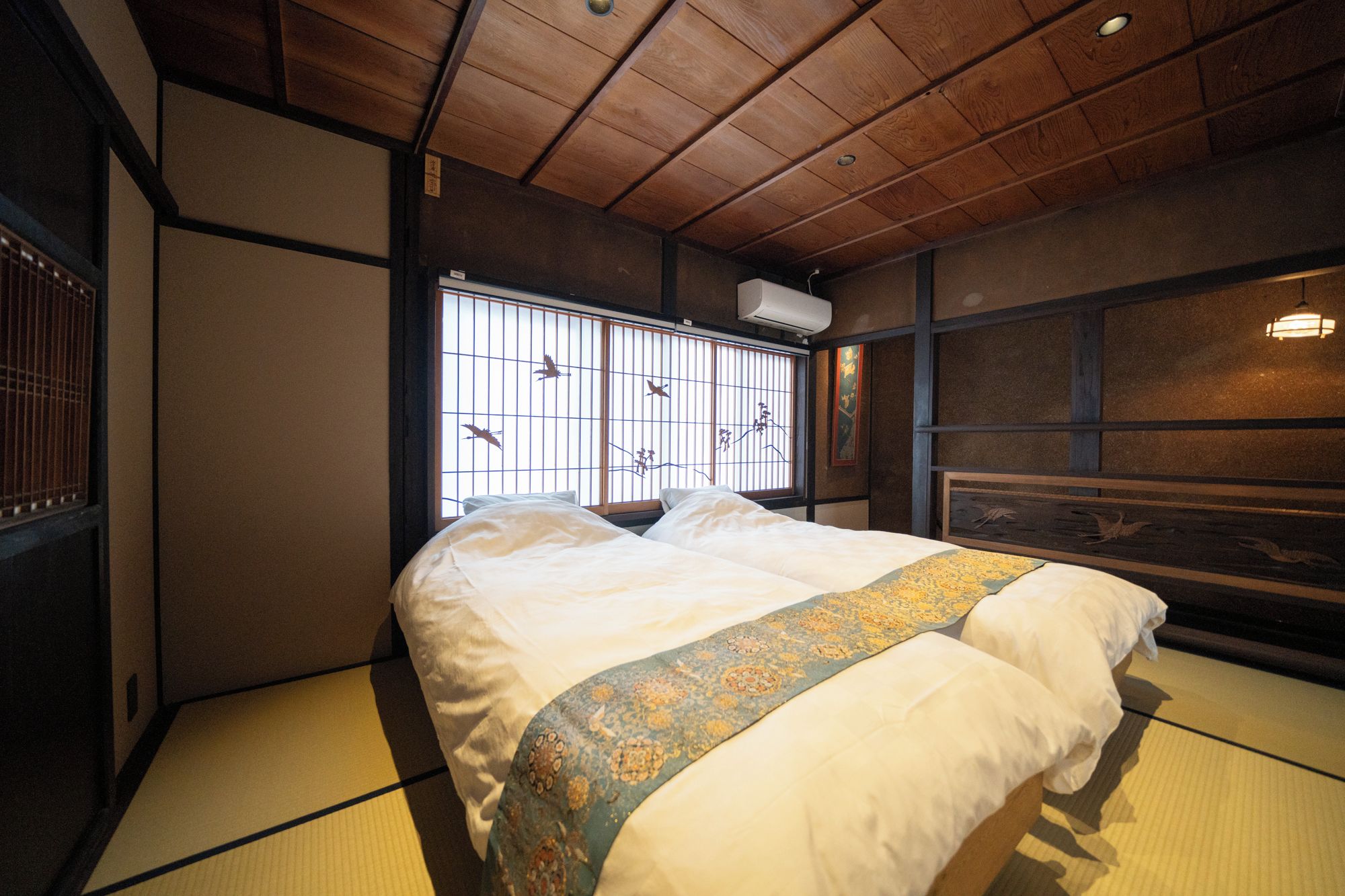A traditional Japanese room with tatami flooring, a bed with a patterned bedspread, a window with a view of birds, and a wooden ceiling.