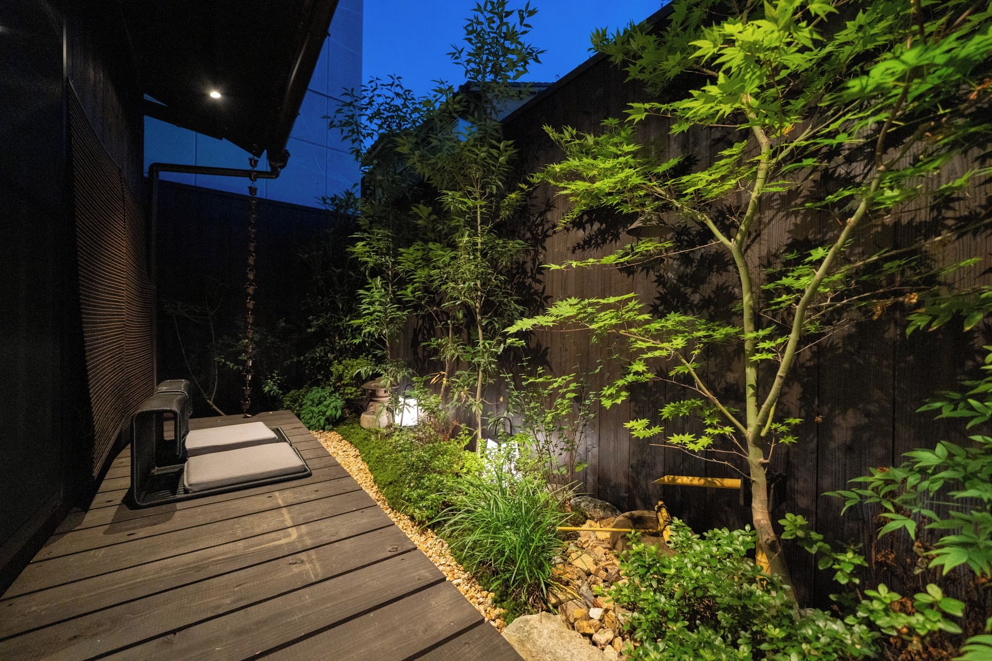 A wooden deck with a stone pathway, surrounded by greenery and a dark wooden wall.