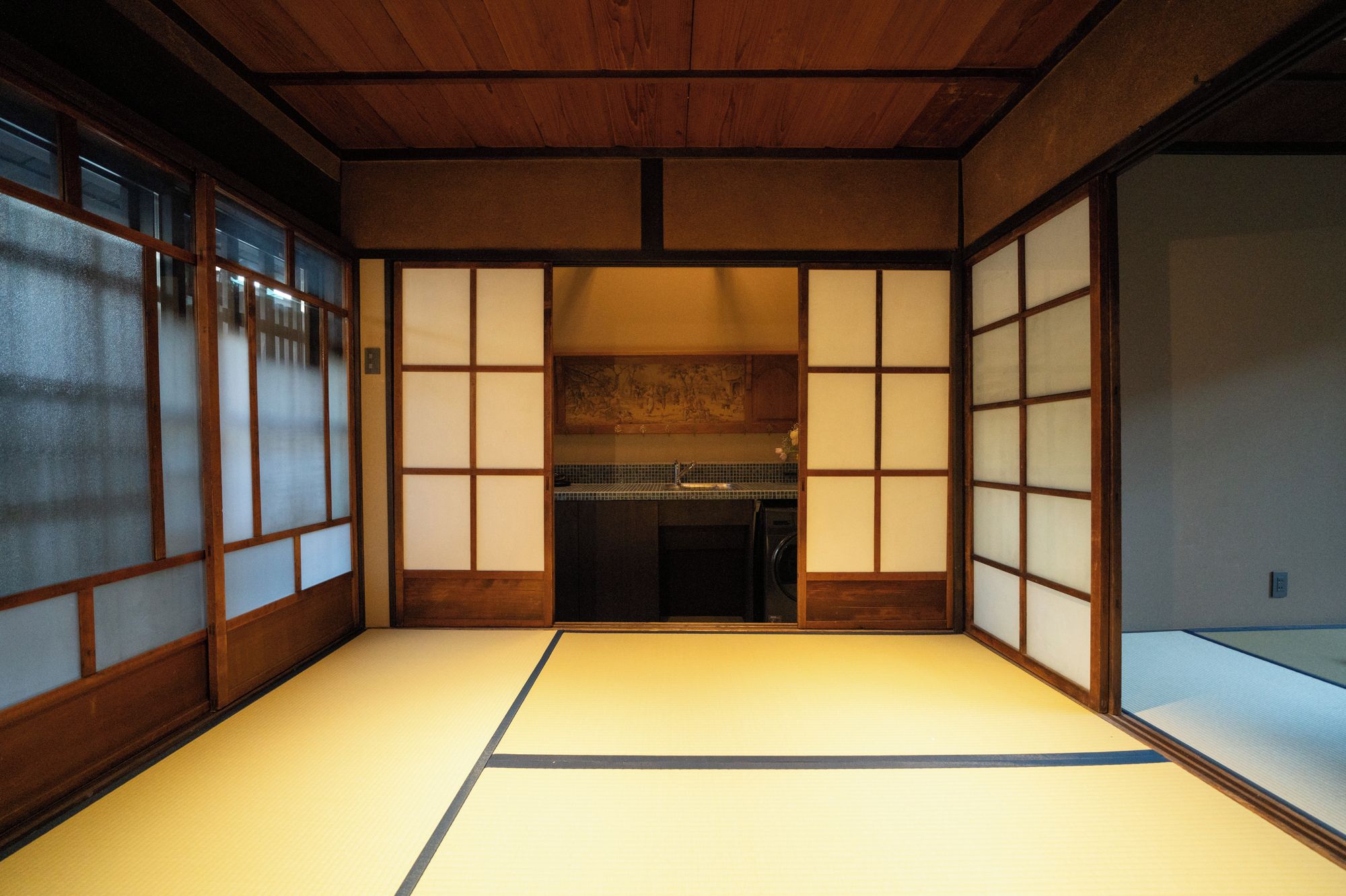 A traditional Japanese room with tatami flooring, sliding doors, and a view of a kitchen area.