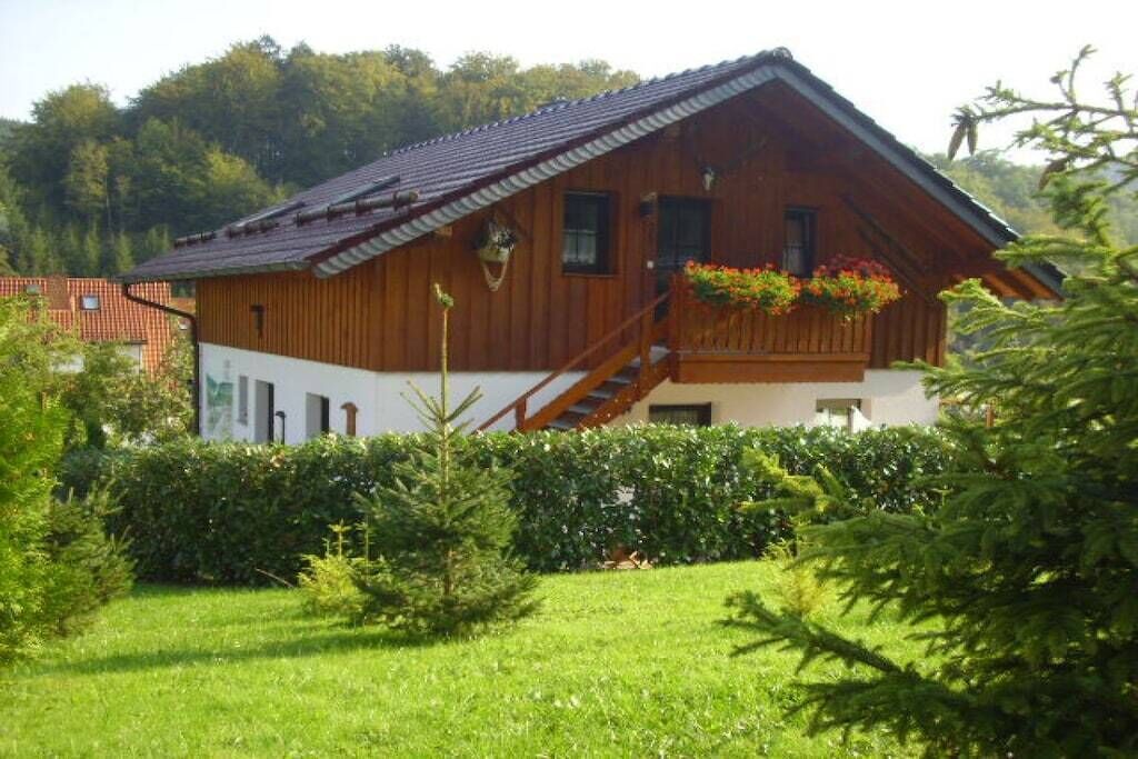 Una casa de madera con techo inclinado, una escalera que conduce a la entrada y un jardín con follaje y flores.