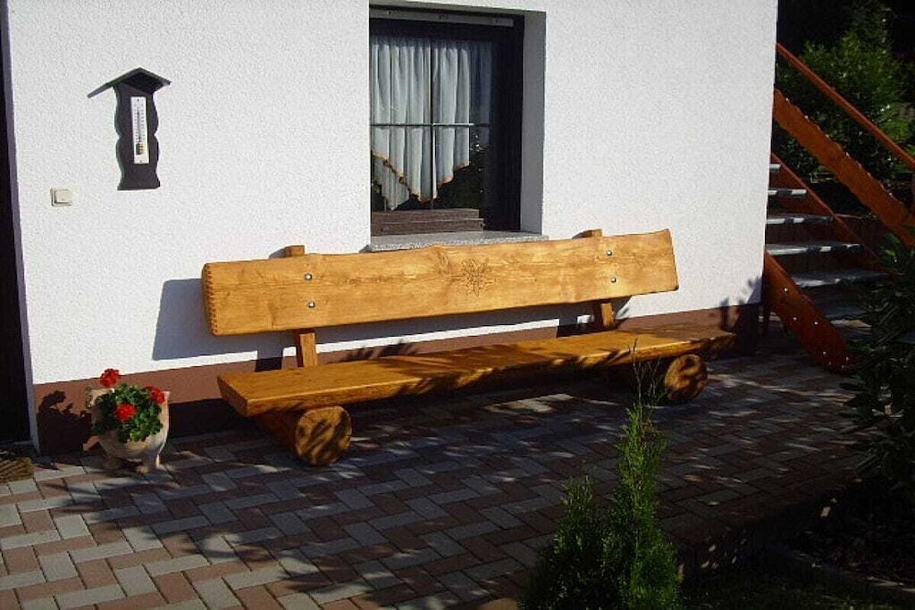 Un banco de madera frente a un edificio con pared blanca, una pequeña ventana con cortinas y una maceta con flores rojas.