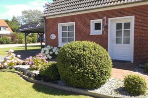 A brick house with two white doors, a covered porch, and a well-maintained garden with flowers and shrubs. - Holiday apartment with 1 bedroom (Krummhörn)