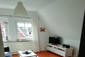 Une pièce avec un meuble blanc pour la télévision, un téléviseur à écran plat, un lampadaire blanc au plafond et une fenêtre donnant sur un bâtiment à l’extérieur.