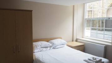 A hotel room with a bed, a wooden wardrobe, a bedside table, and a window with a view of the outside.