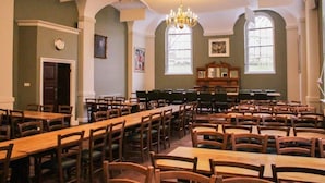 A spacious room with multiple wooden tables and chairs, a chandelier, and large windows.