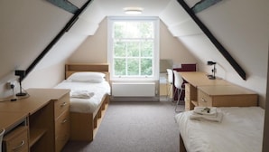 Un pequeño dormitorio bajo los tejados, bien iluminado, con dos camas, un escritorio y una ventana que proporciona luz natural.