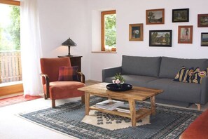 Ein Wohnzimmer mit einer Couch, einem Sessel, einem Couchtisch und einem Fenster mit Blick ins Grüne.