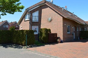 A two-story brick house with a sloped roof, a sunburst decoration on the facade, a gated driveway, and a well-maintained hedge. - Holiday home with 2 bedrooms (Greetsiel)