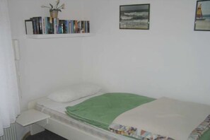 Une chambre avec un lit simple, une couverture verte, une table de chevet, une étagère avec des livres et une petite plante, ainsi que deux tableaux encadrés accrochés au mur.