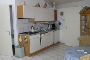 A compact kitchen with white cabinets, a stove, and a sink. There is a small trash bin and a wicker chair in the dining area. - in the Strandslag 103 holiday park in Julianadorp (Julianadorp)