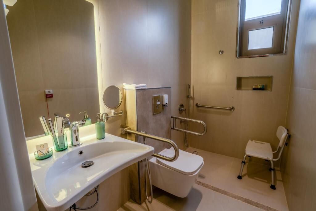 A bathroom with a white sink, mirror, toilet, and a wheelchair-accessible chair.