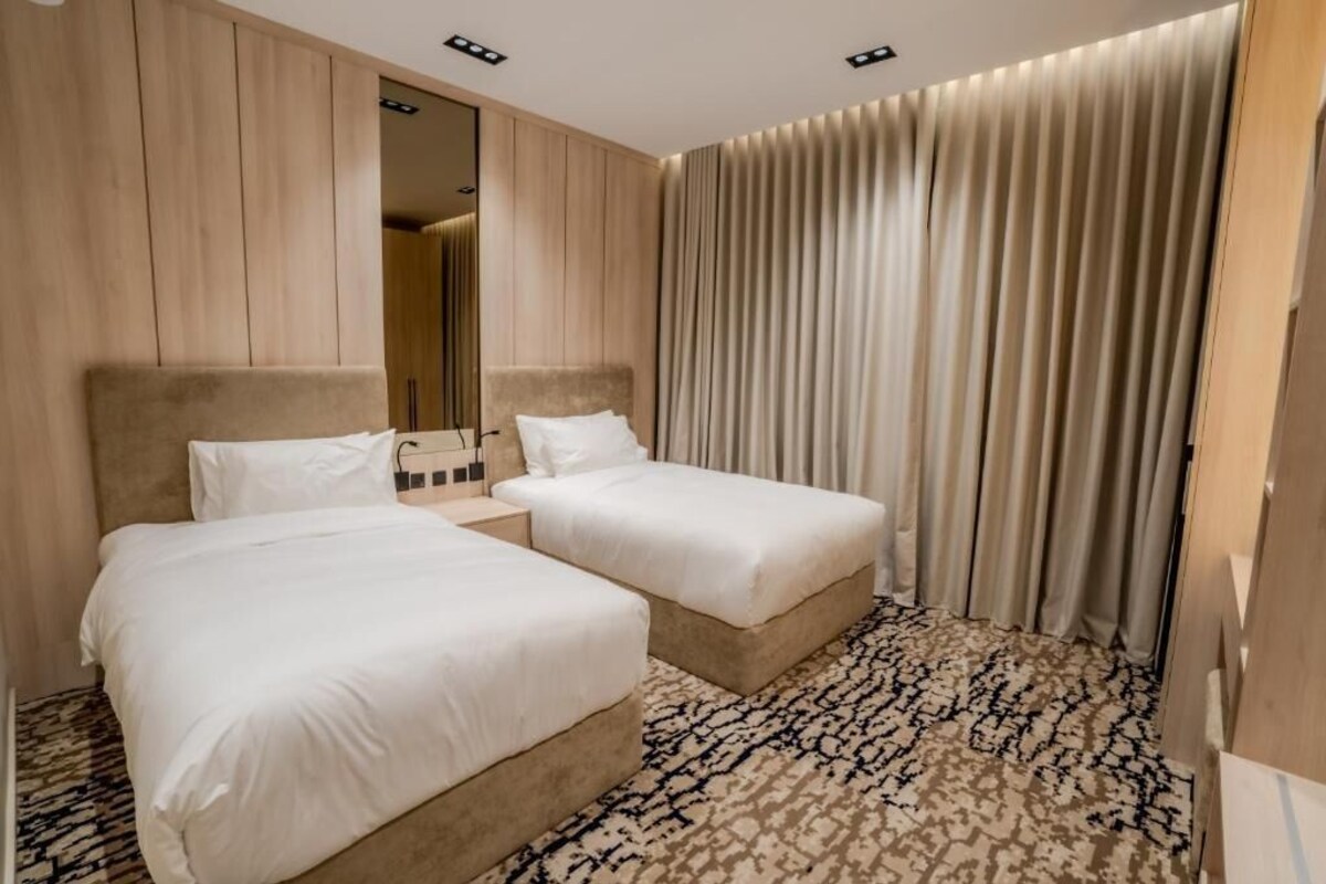 Een hotelkamer met twee bedden, een kamerbreed tapijt met dessin, houten paneelwanden en een groot raam met gordijnen.