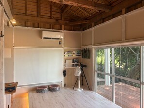 A room with a wooden ceiling, a white air conditioning unit, a tripod with a camera, and a large window with a view of trees. - Keshiki Chosan｜Camping, BBQ & Bonfire (富津市萩生)