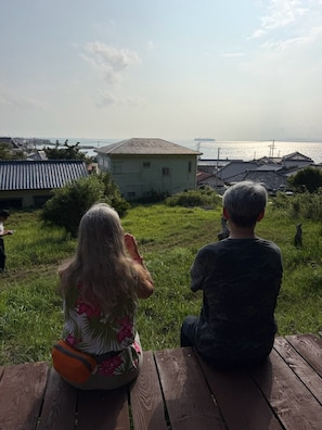 Two individuals seated on a wooden bench overlooking a grassy area with a view of a body of water and buildings in the distance. - Keshiki Chosan｜Camping, BBQ & Bonfire (富津市萩生)