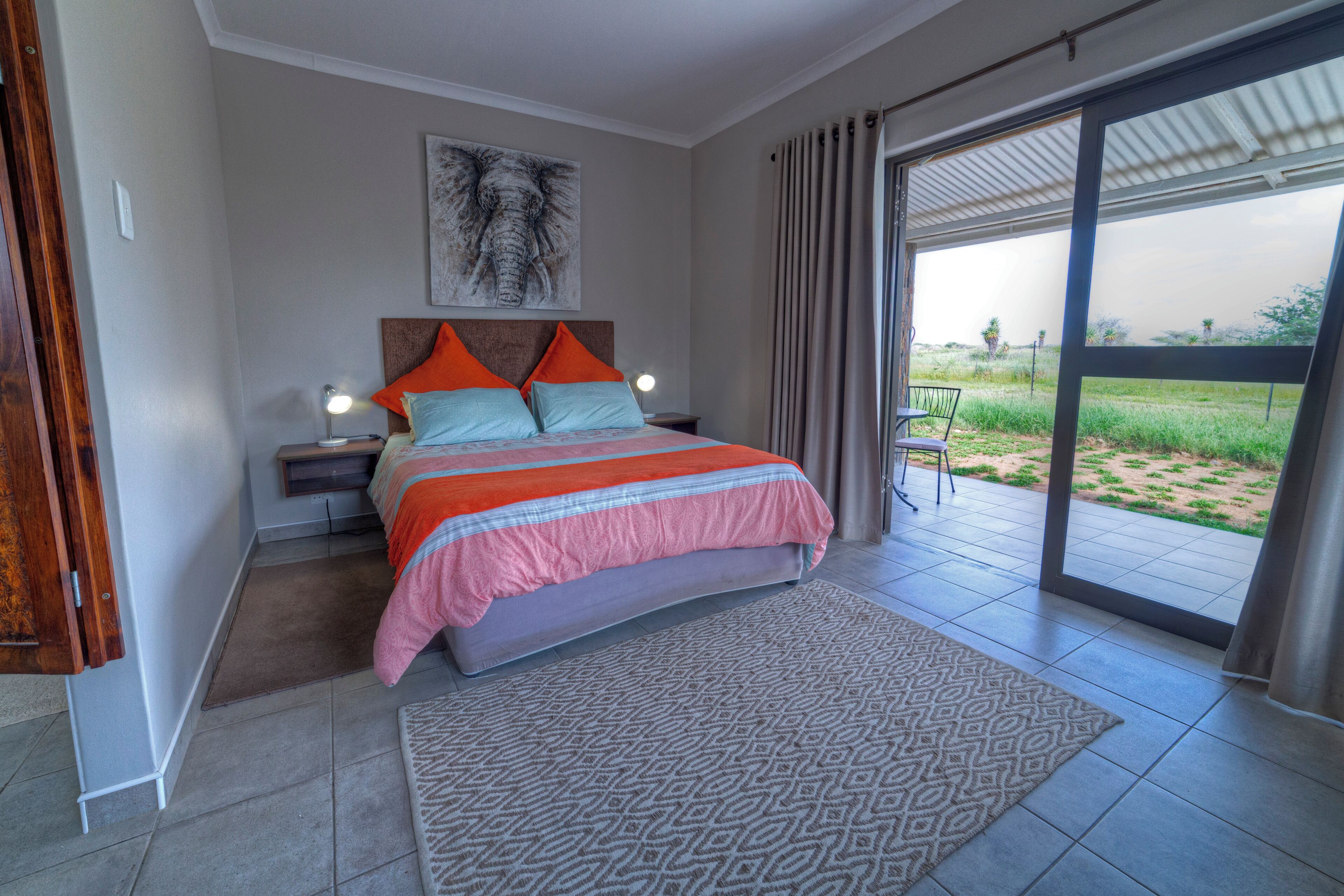 A bedroom with a bed, nightstands, a lamp, a large window, and a view of the outdoors.
