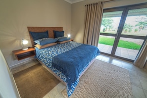 Un dormitorio con cama, mesitas de noche, lámpara, ventana con cortinas y vista a zonas verdes.