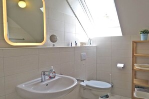 Een badkamer met een witte wastafel, toilet en een spiegel met verlichting. Boven het toilet bevindt zich een dakraam.