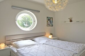 Een slaapkamer met een bed, een rond raam, een wandplank en een plafondlamp.