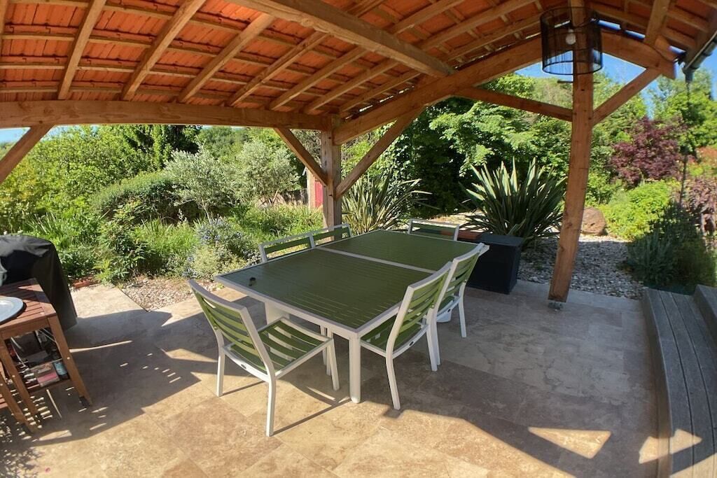 An outdoor patio with a table and chairs, surrounded by greenery and a wooden pergola.
