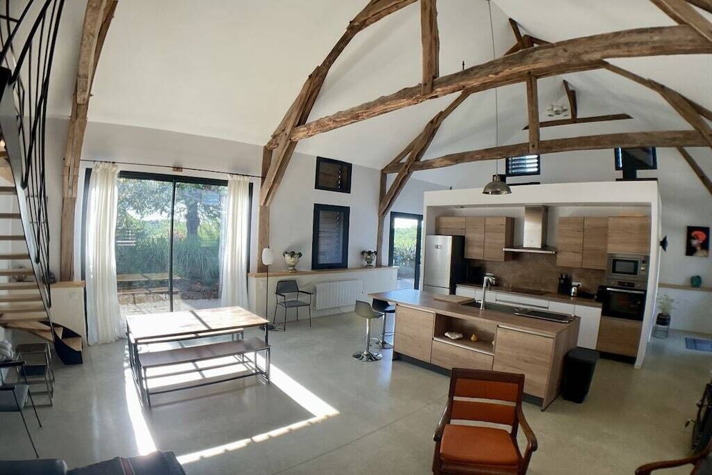 Un vaste espace de vie comprenant un coin repas avec une table, une cuisine équipée d’appareils modernes et un escalier avec des poutres apparentes.