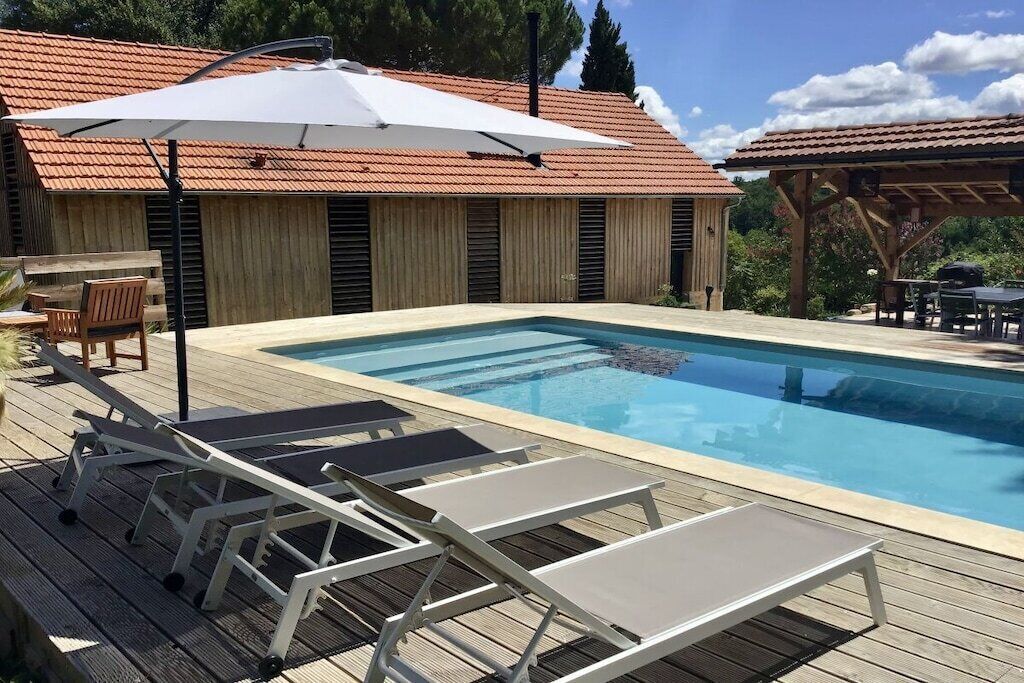 En swimmingpool med solstolar och ett parasoll, ett trädäck och en byggnad med ett tak täckt av tegelpannor.