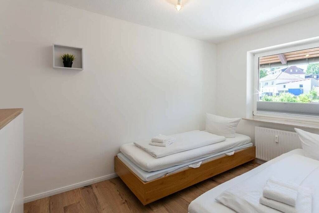 A bedroom with a bed, a small shelf with a plant, and a window with a view of buildings.