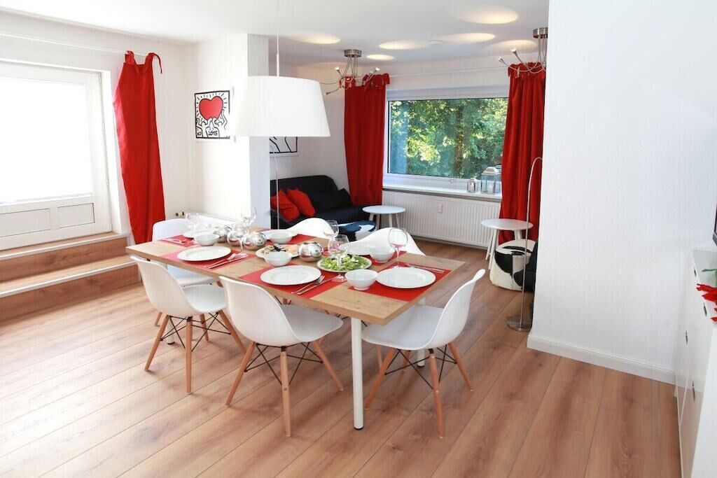 Un coin repas avec une table en bois dressée pour quatre personnes, des chaises blanches et un chemin de table rouge.