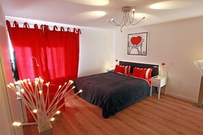 Een slaapkamer met een bed, rode gordijnen, een nachtkastje, een wanddecoratie en een kroonluchter.