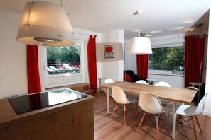 Een eetruimte met een houten tafel en witte stoelen, een groot raam met rode gordijnen, en een plafondlamp.